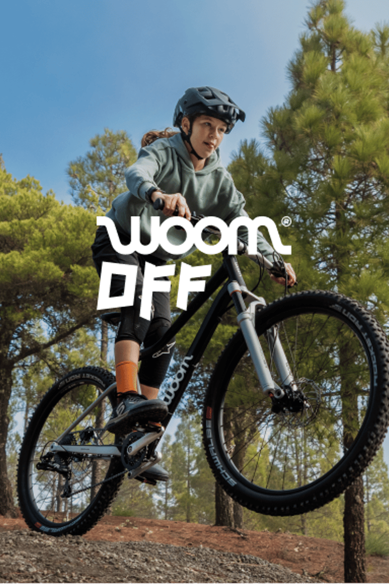 An image of a girl wearing a helmet and beginning a jump on a black woom OFF bike in a forested area.