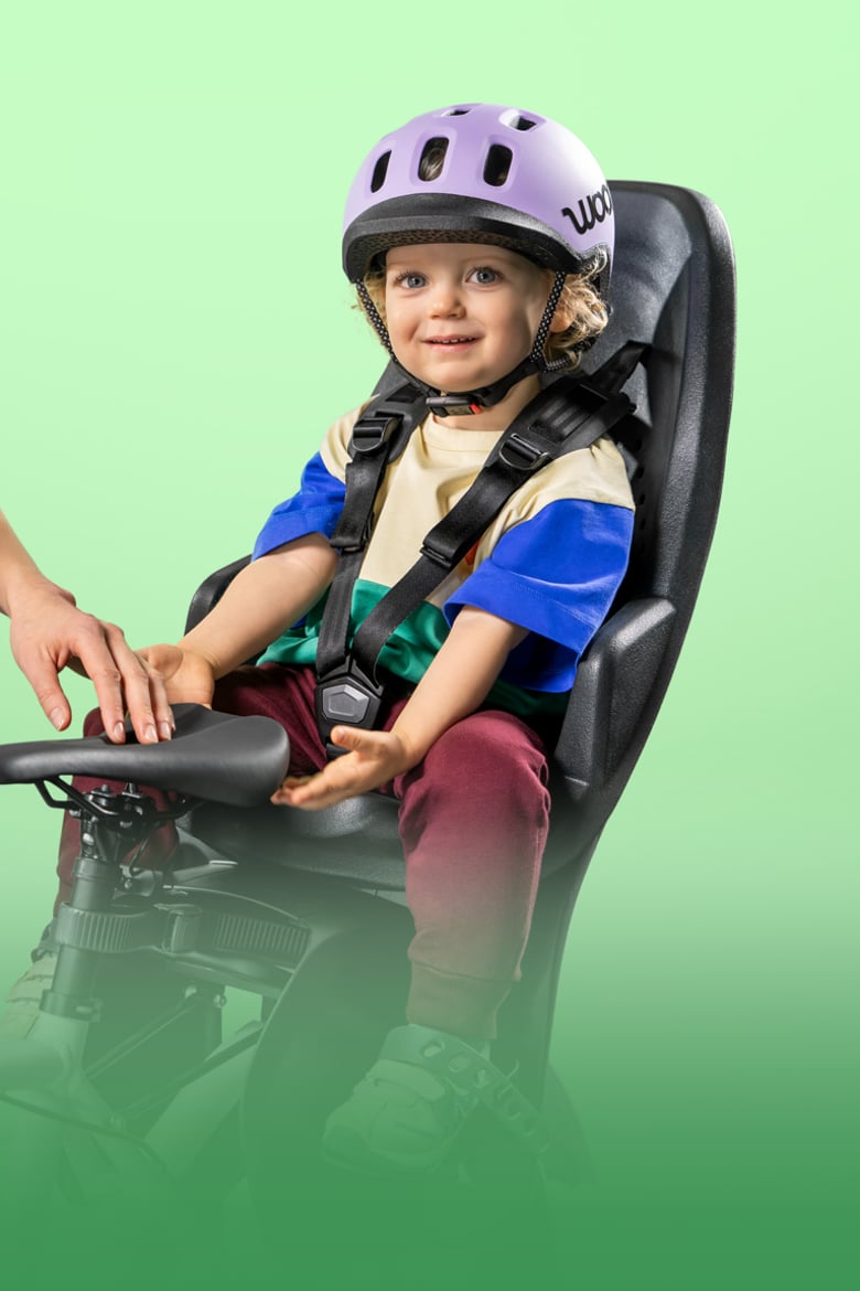 Lächelndes Kleinkind mit lilafarbenem READY Kinderhelm auf dem Kopf sitzt im Fahrradsitz vor grünem Hintergrund.