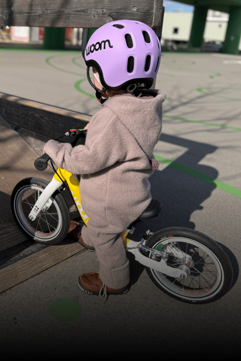 Ein Kleinkind mit lila READY Radhelm und Wollwalkanzug steht auf einem gelben woom GO Laufrad in 12 Zoll.