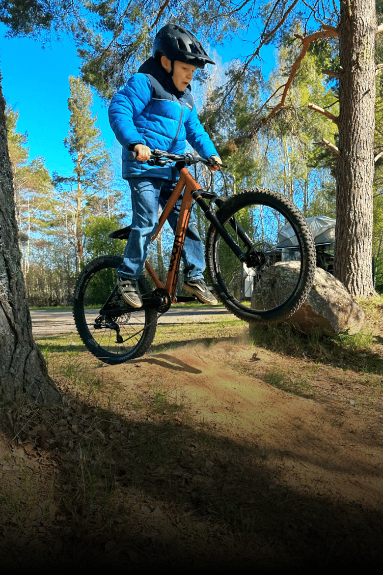 A boy is riding a copper-colored woom OFF AIR with the front wheel off the ground in a woodland setting.