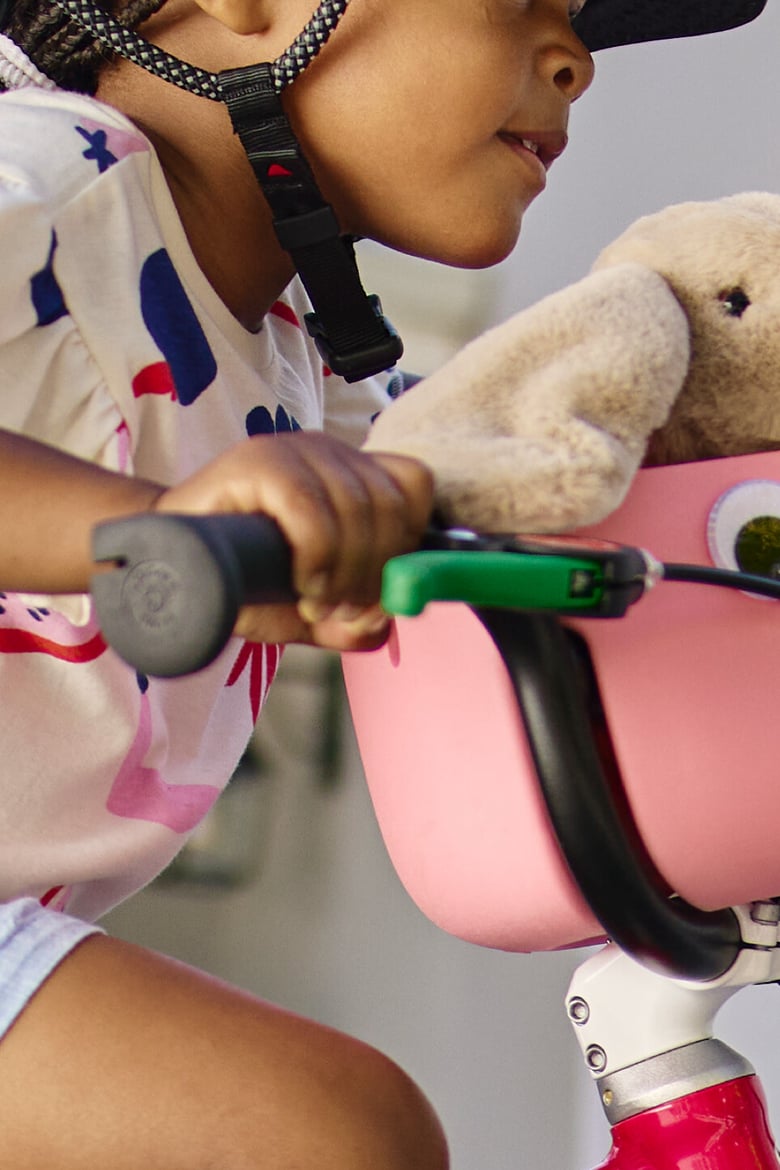 A young girl wearing a woom READY helmet rides a woom red GO bike with attached marshmallow pink POP basket holding a stuffed rabbit toy.