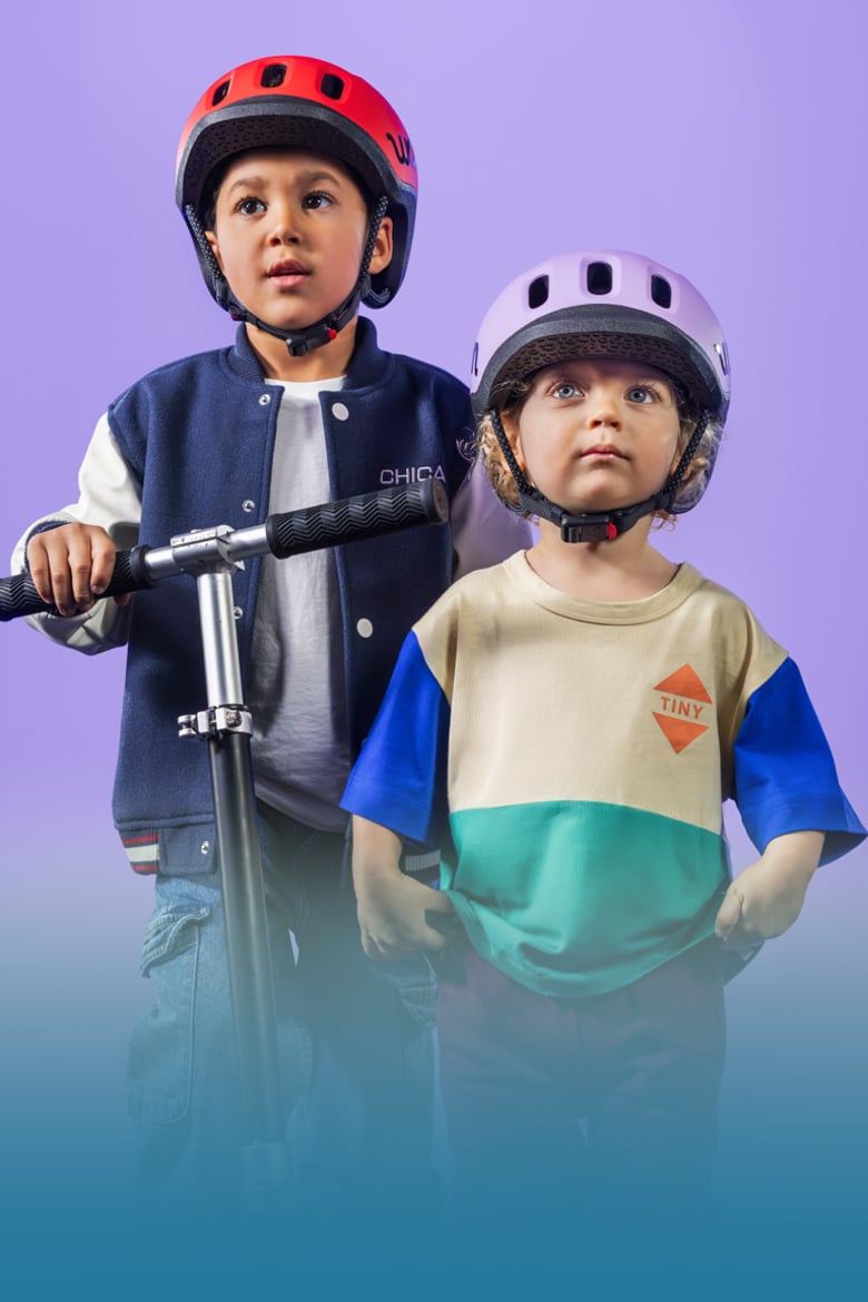 Zwei Kinder mit READY Kinderhelmen von woom auf den Köpfen und einem Scooter in der Hand blicken in die Ferne.
