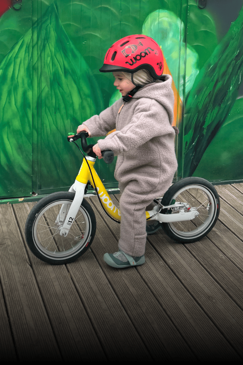 Auf einem gelben woom Laufrad in 12 Zoll rollt ein Kleinkind mit rotem READY Radhelm und Wollwalkanzug.