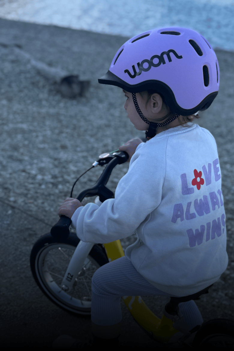 Un petit enfant portant un casque READY violet est assis sur une draisienne jaune de woom.