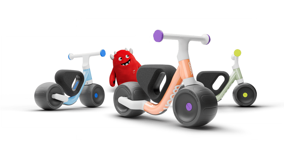 The woom WOW toddler balance bike lineup showcased in blue, orange, and green colorways, featuring the red soft toy Buddy.