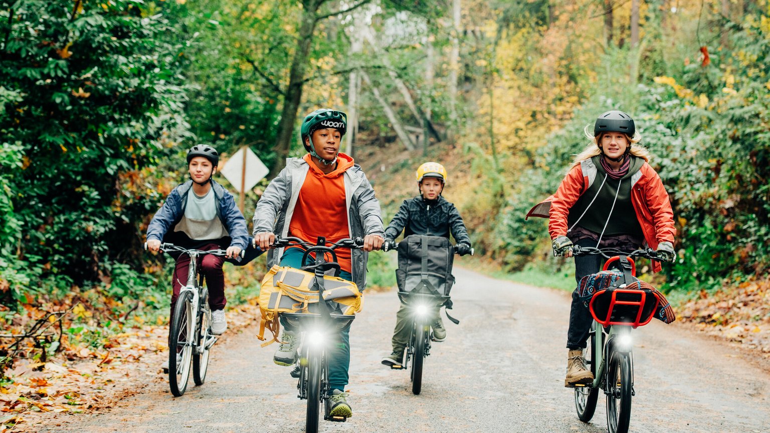 Quatre enfants roulent sur leurs vélos woom NOW et woom ORIGINAL de 26 pouces sur une route à travers la forêt.