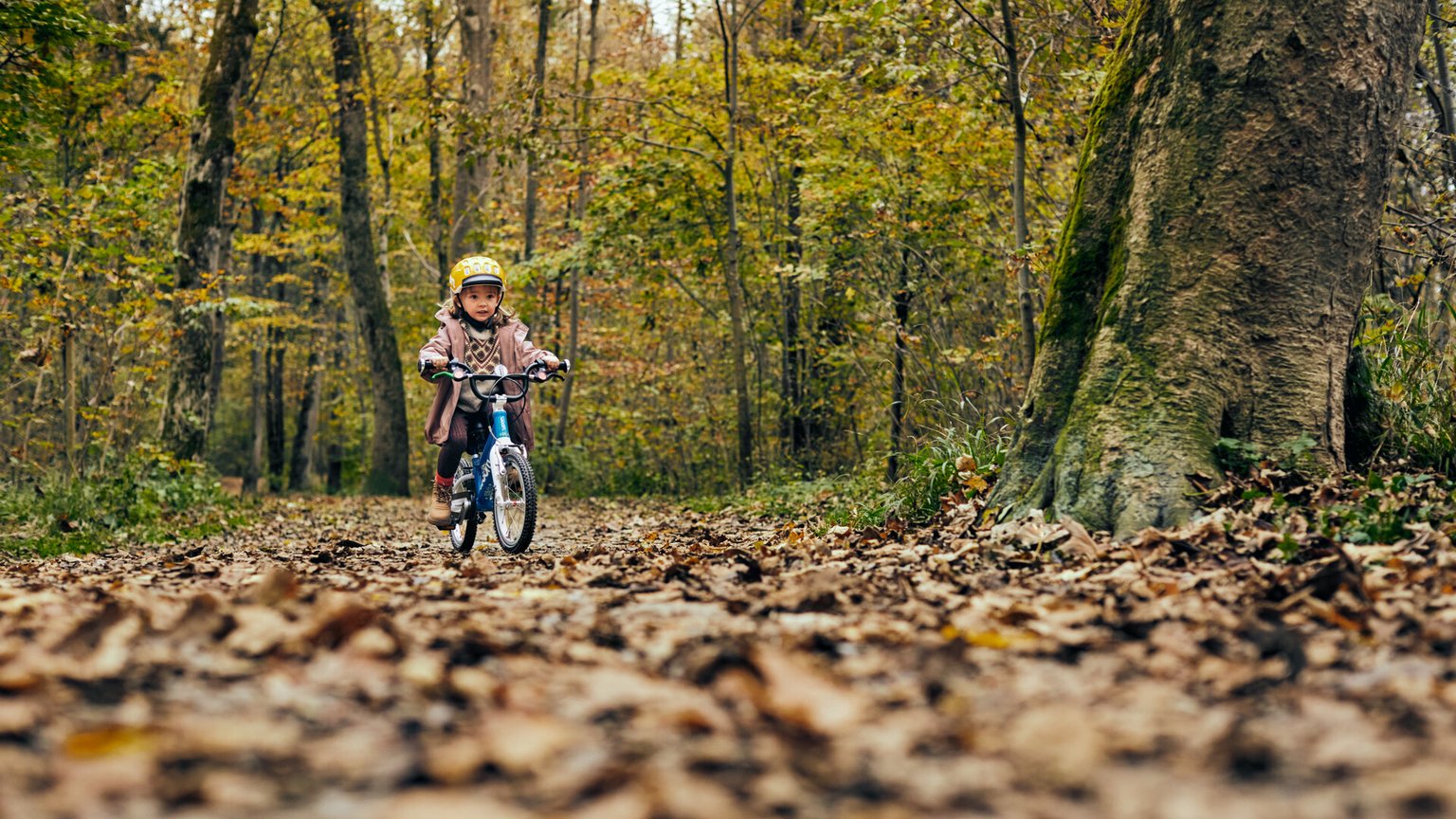 Ein kleines Mädchen mit gelbem woom Helm fährt auf einem blauen woom ORIGINAL 2 Kinderfahrrad in 14 Zoll durch den Wald.