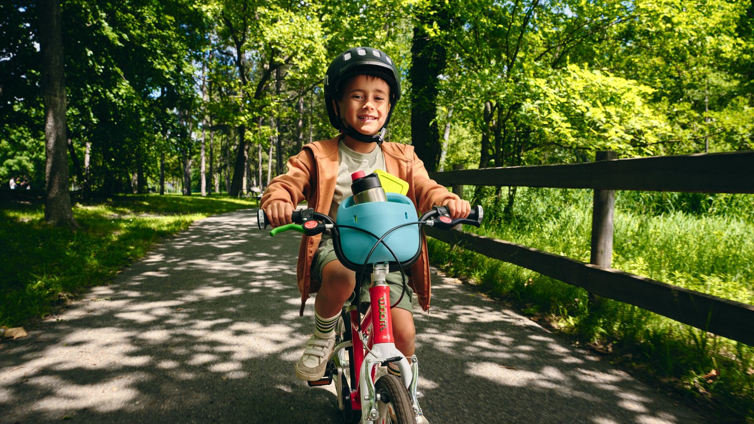 Photo d’un petit garçon vu de face sur son vélo woom rouge et blanc avec un panier à vélo bleu ciel sur le guidon, dans lequel se trouve une gourde.