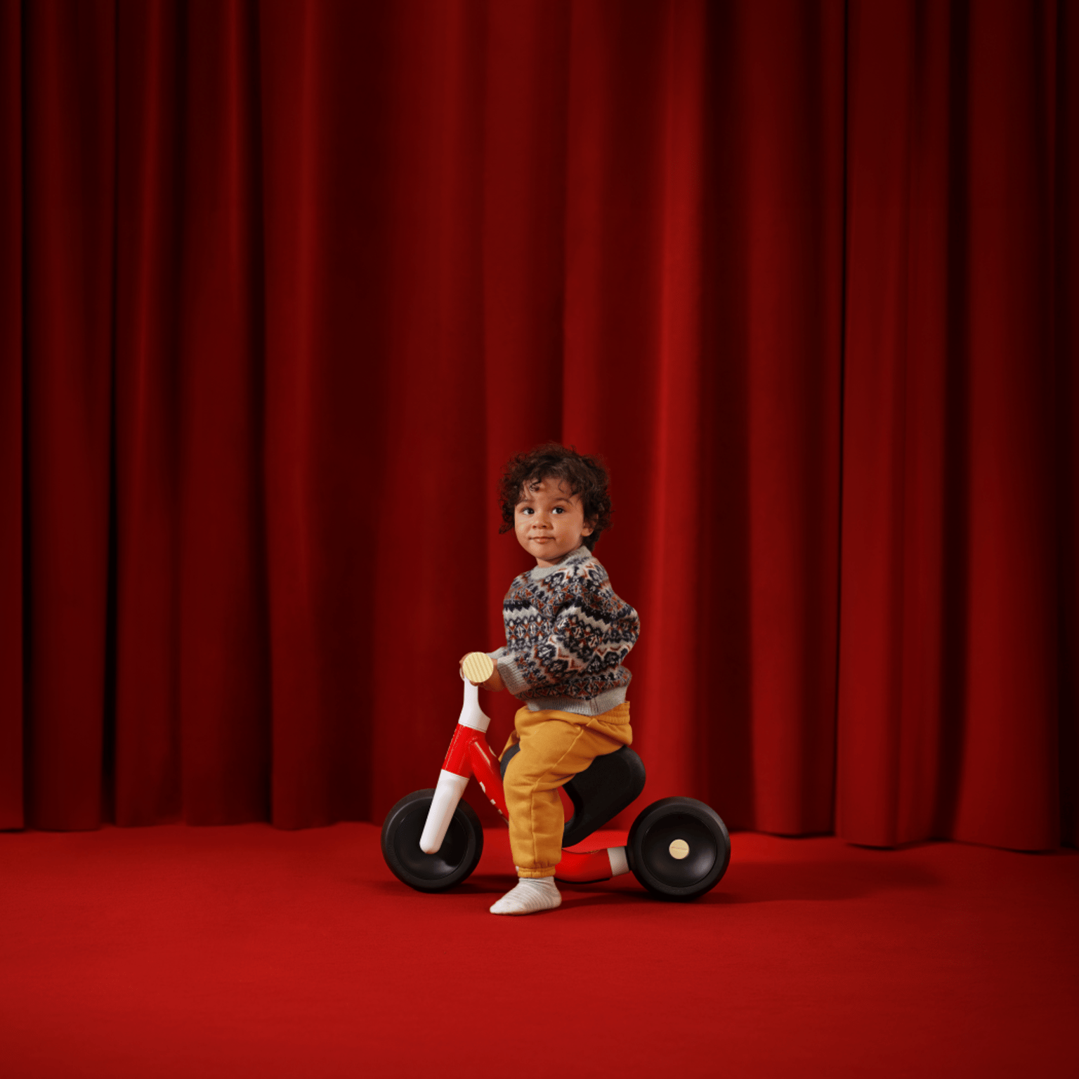 A young boy sits in profile atop a new red woom WOW in front of a red velvet curtain and on top of a red carpet.