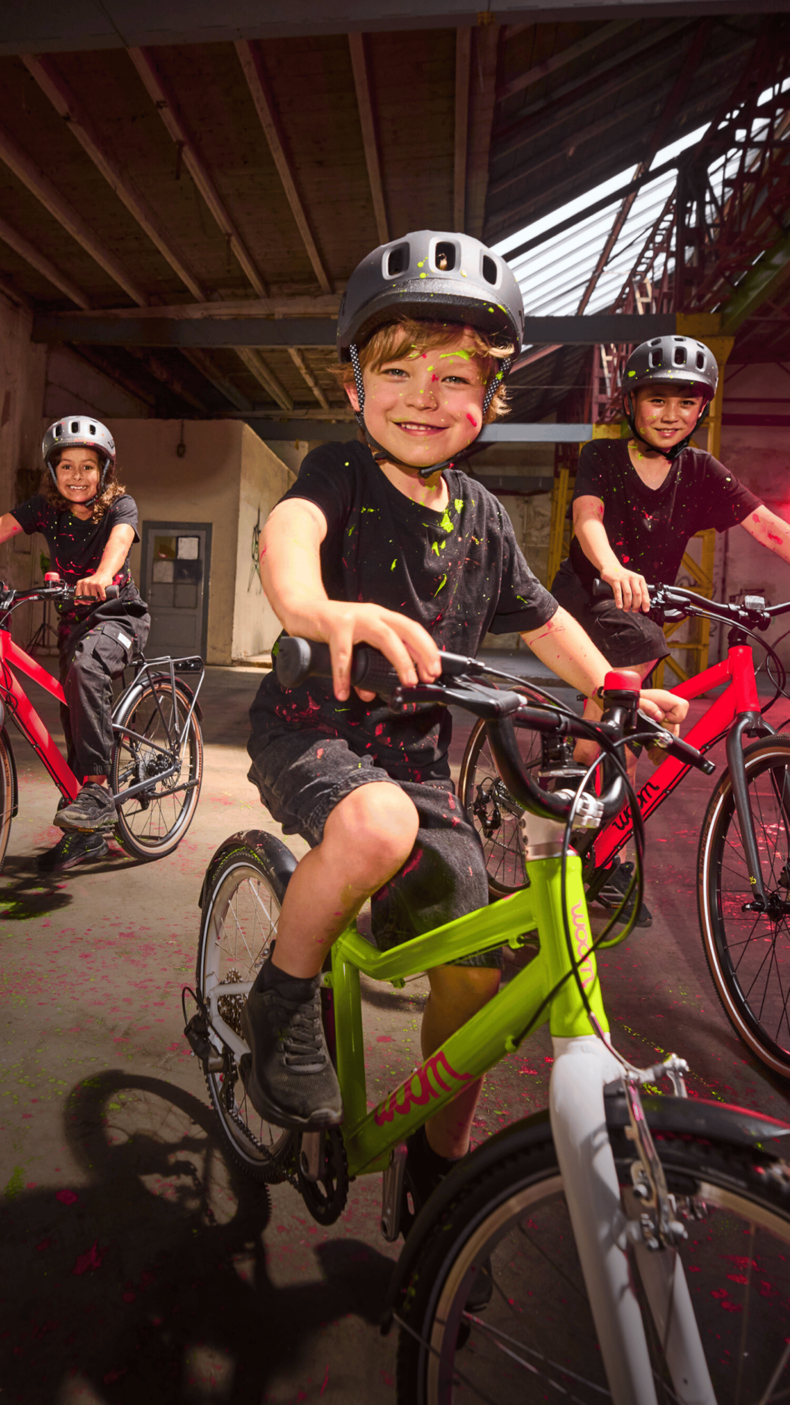 Drei Kinder mit Helmen auf neonfarbenen woom Bikes in einer alten Industriehalle mit bunten Farbspritzern.