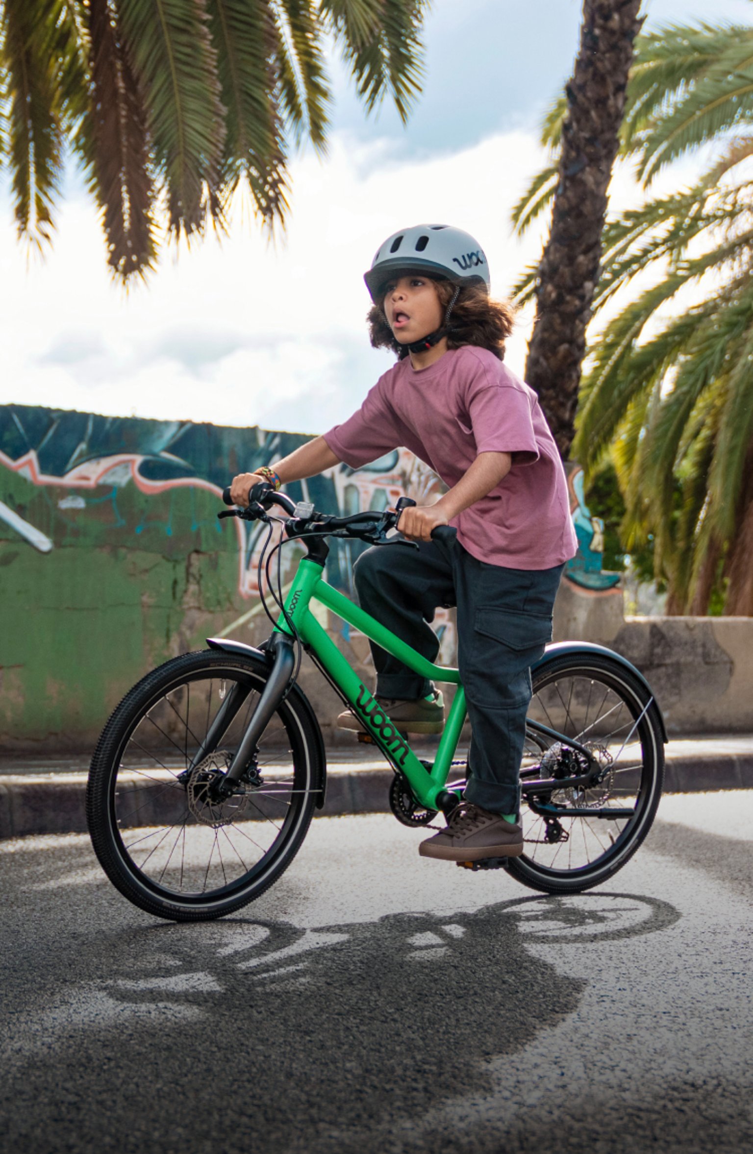 Un enfant portant un casque enfant READY roule sur son vélo woom EXPLORE vert devant un mur graffé.