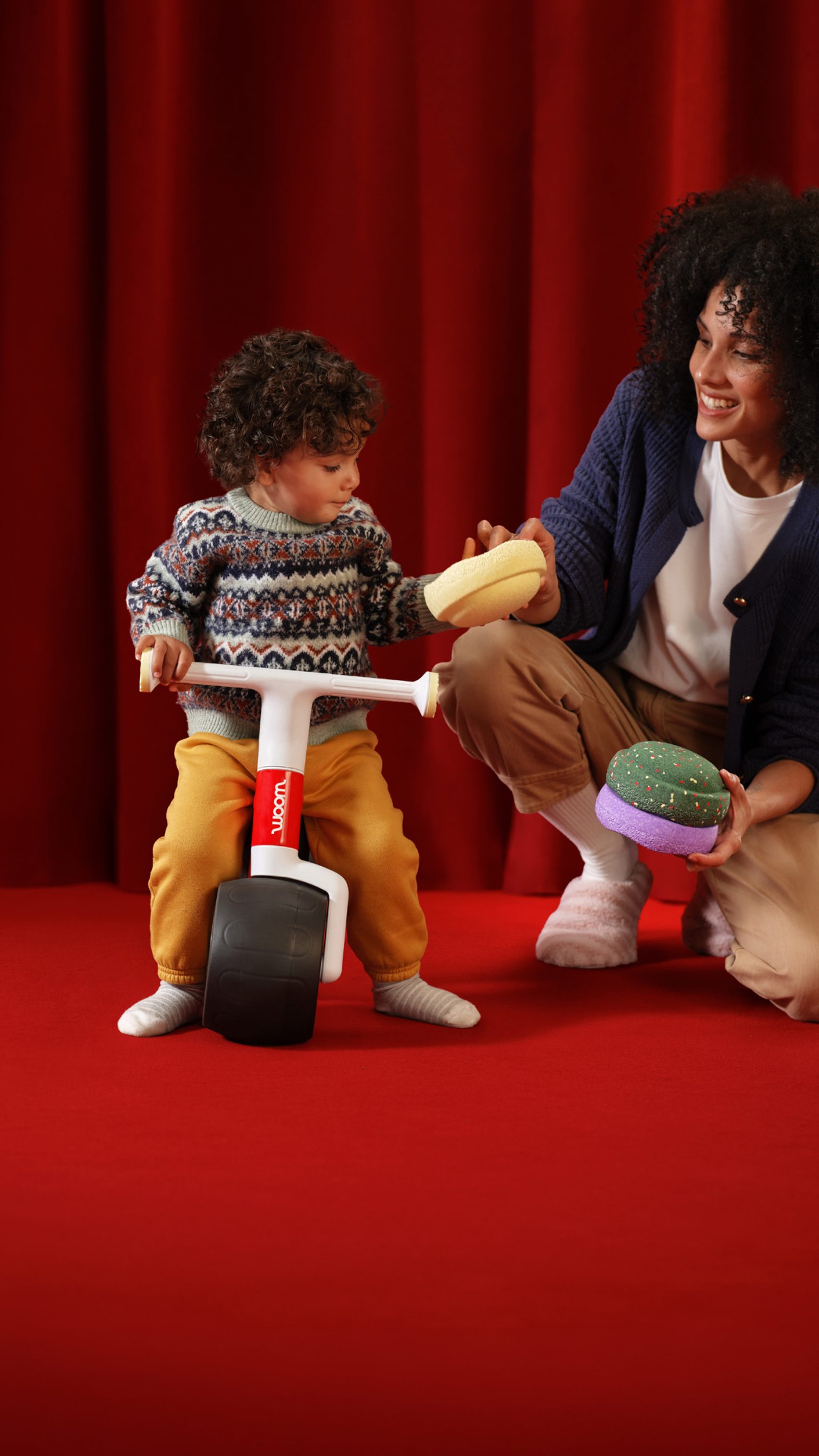 A toddler sitting on the red woom WOW Seasonal Colour Edition balance bike on a red stage, while a woman cheers in the background near scattered exclusive Stapelstein® Original Minis.