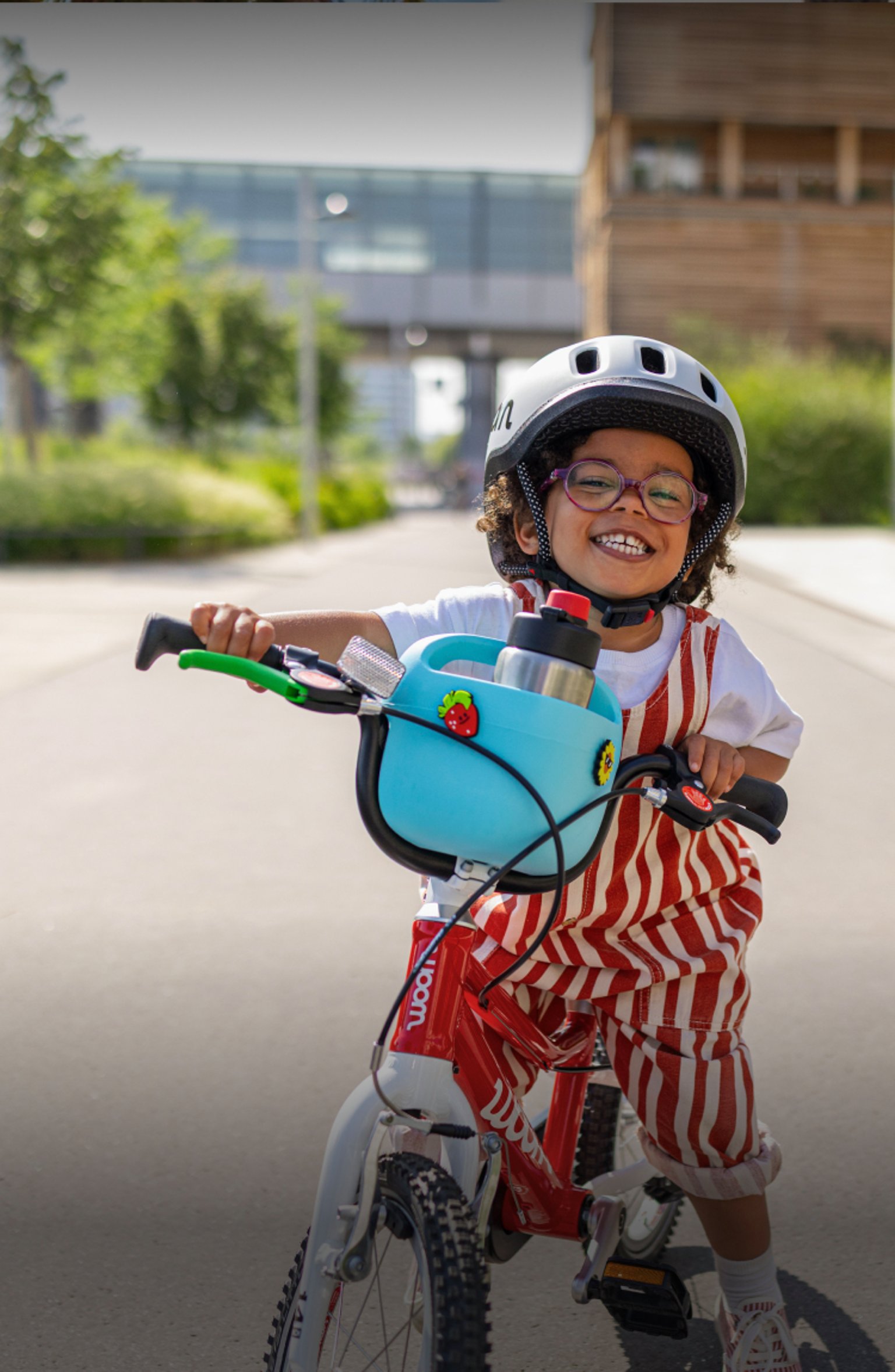 Un enfant souriant avec un casque enfant READY roule sur un vélo woom GO rouge équipé d’un panier POP bleu par temps ensoleillé.