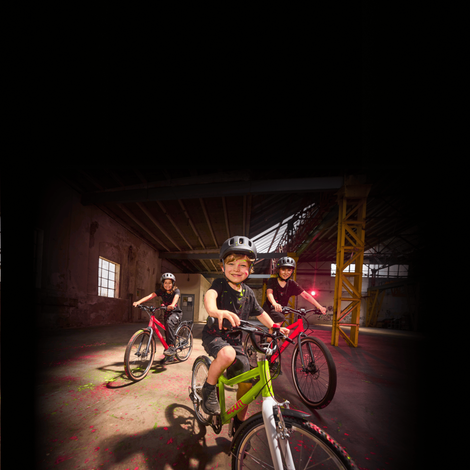 A photo of three kids riding their bikes through a paint-splattered warehouse, highlighting the electric new colors featured on each bike: neon lime woom GO and neon pink woom EXPLORE.