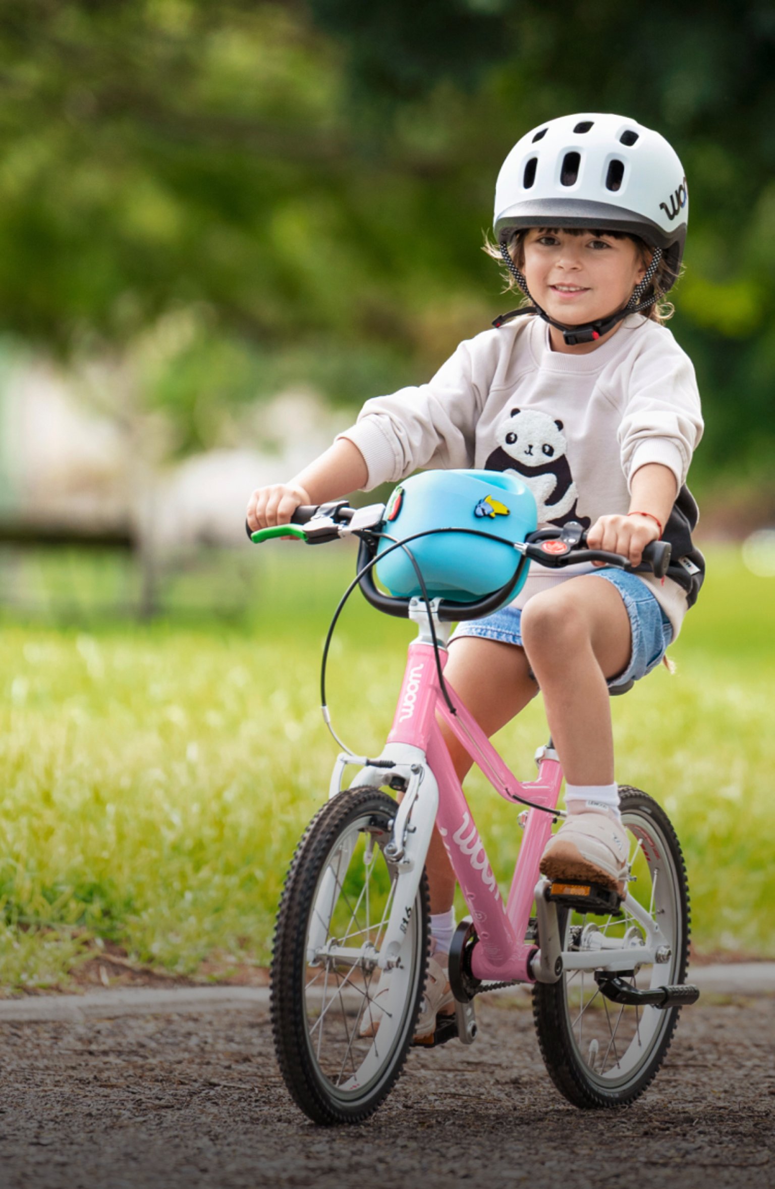 Un enfant avec un casque READY gris pédale sur un vélo woom GO powder pink ; une femme l’encourage, les bras ouverts. 