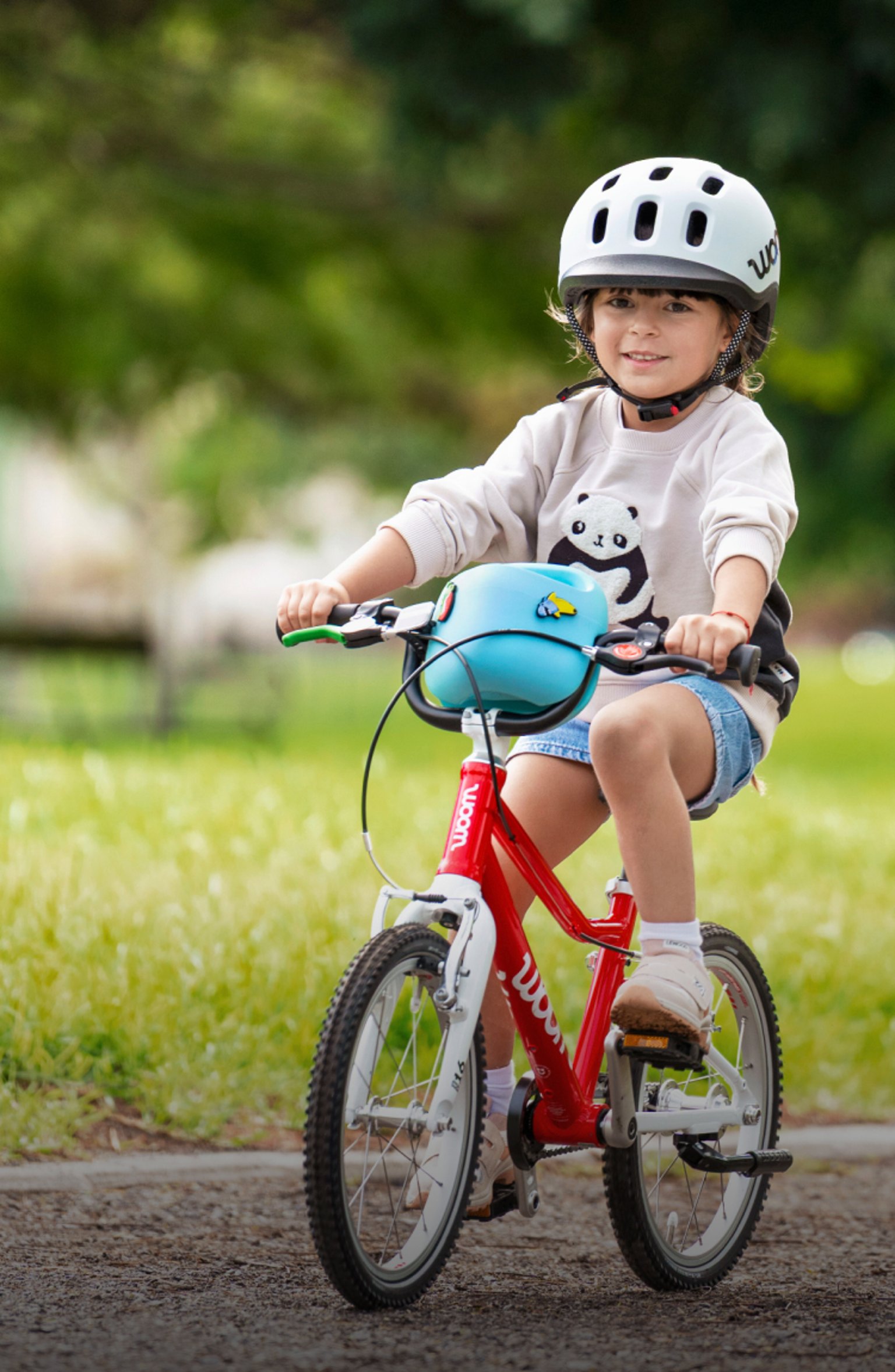 Kind mit woom READY Helm fährt auf einem roten woom GO, während eine Frau im Park zuschaut.