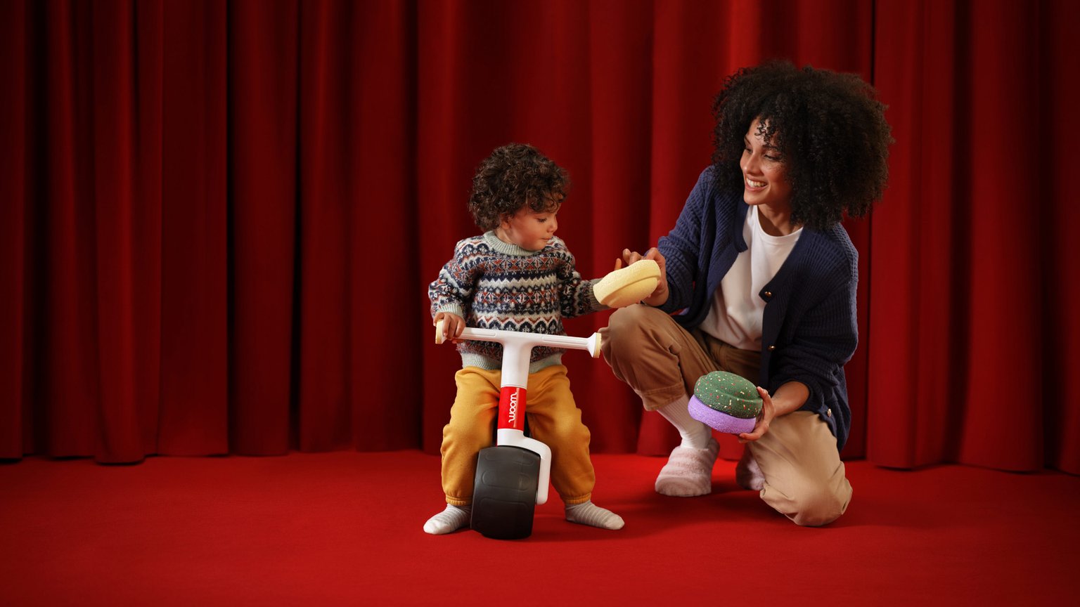 A toddler enjoying the woom red WOW balance bike while playing with the exclusive colorful Stapelstein® Original Minis alongside a smiling adult, set against a festive red backdrop.