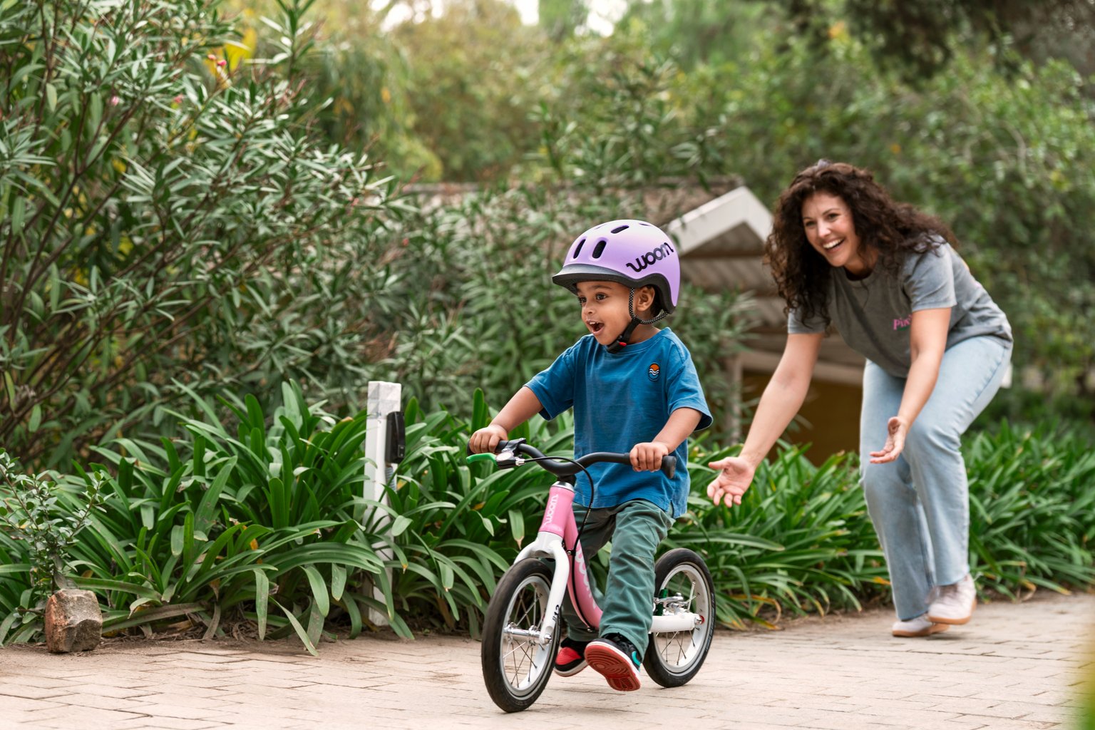 Un enfant avec un casque woom READY violet sur une woom GO 1 en powder pink est encouragé par une femme.