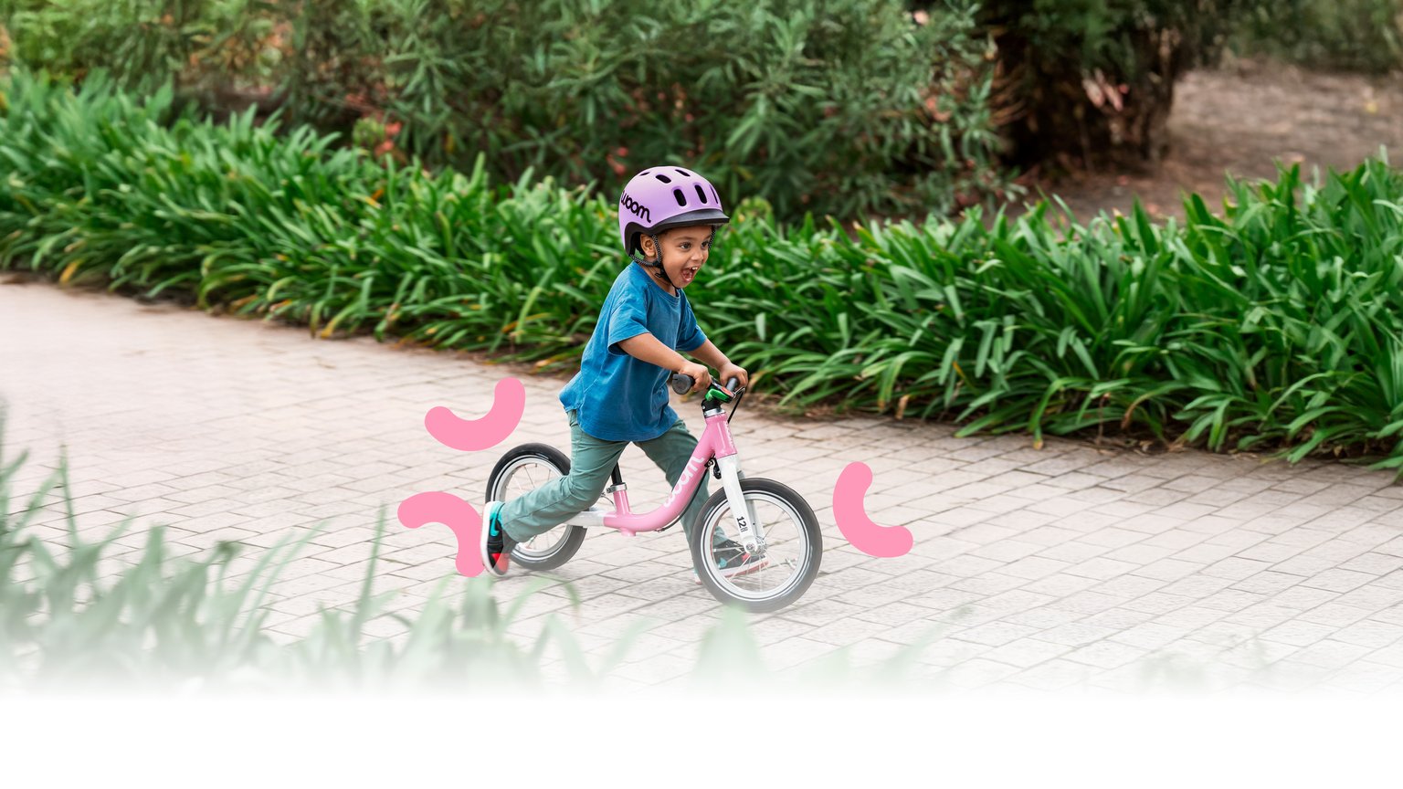 Un enfant portant un casque READY violet roule avec sa woom GO rose sur un chemin pavé dans un cadre vert et fleuri.
