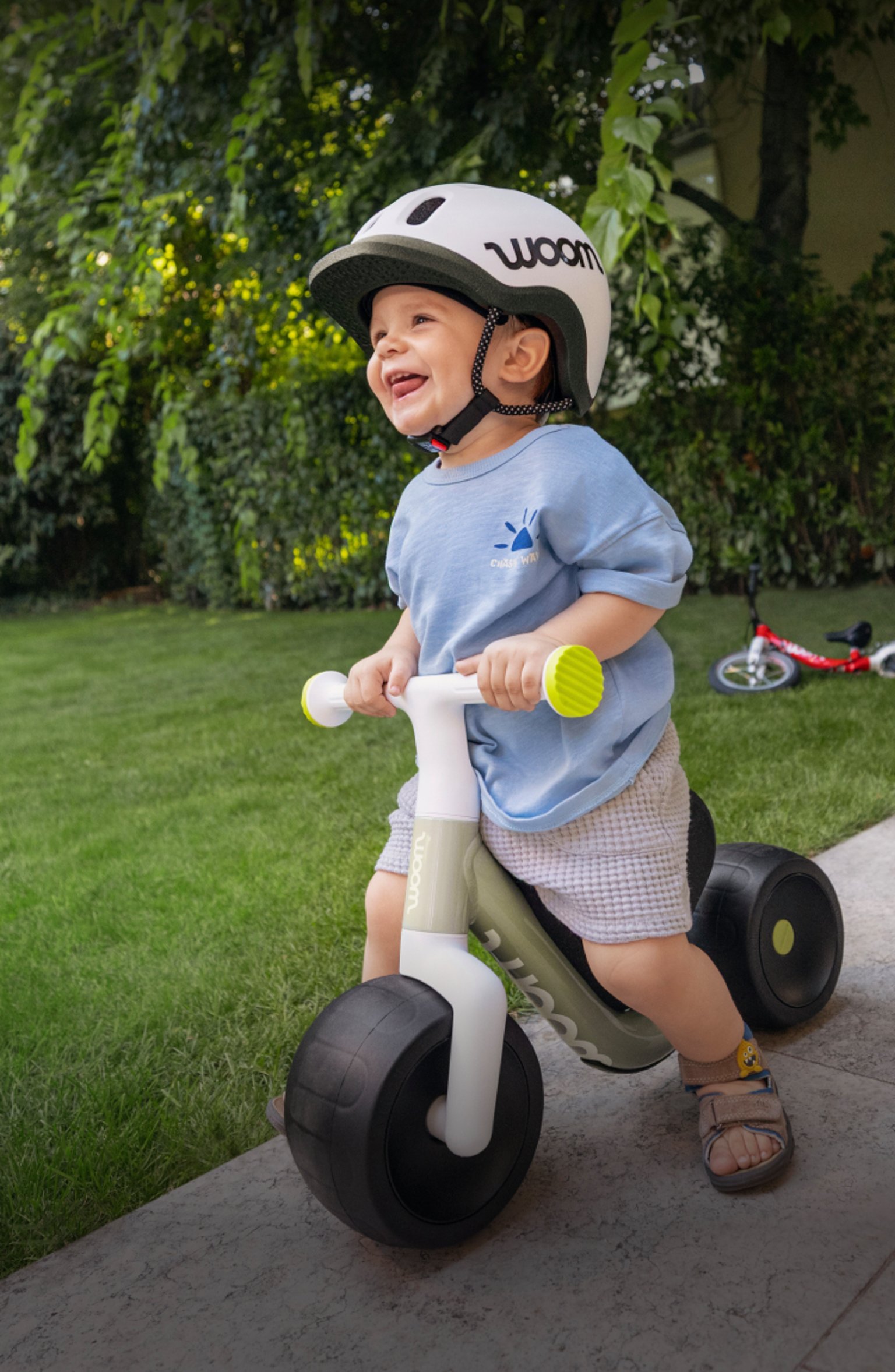 Un enfant souriant portant un casque avec sa draisienne dans un jardin. Bannière publicitaire pour les woom WOW et woom GO.