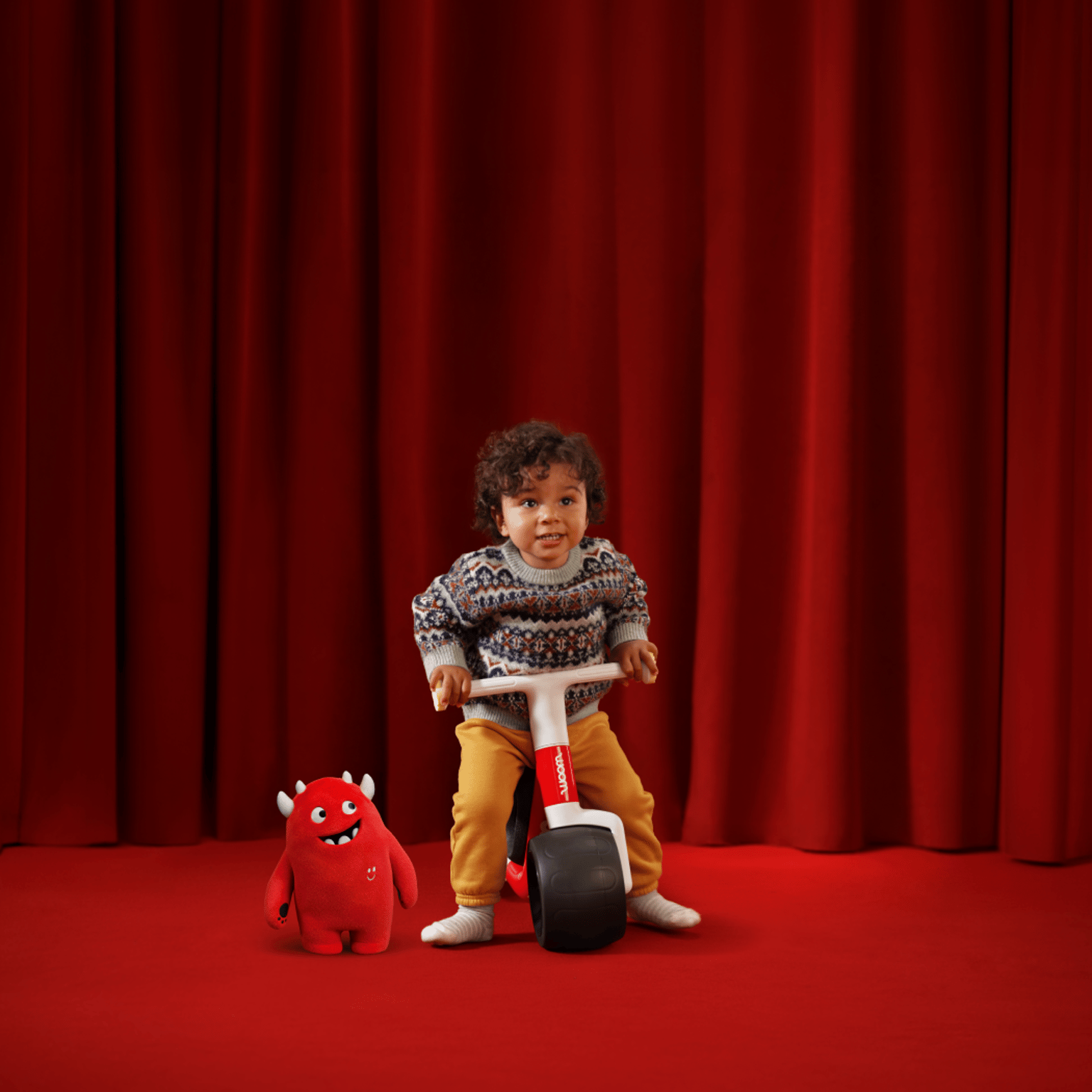 A young boy sits facing the camera atop a new red woom WOW in front of a red velvet curtain and on top of a red carpet. Buddy, the red woom monster mascot, stands to the left of the bike smiling up at the boy.