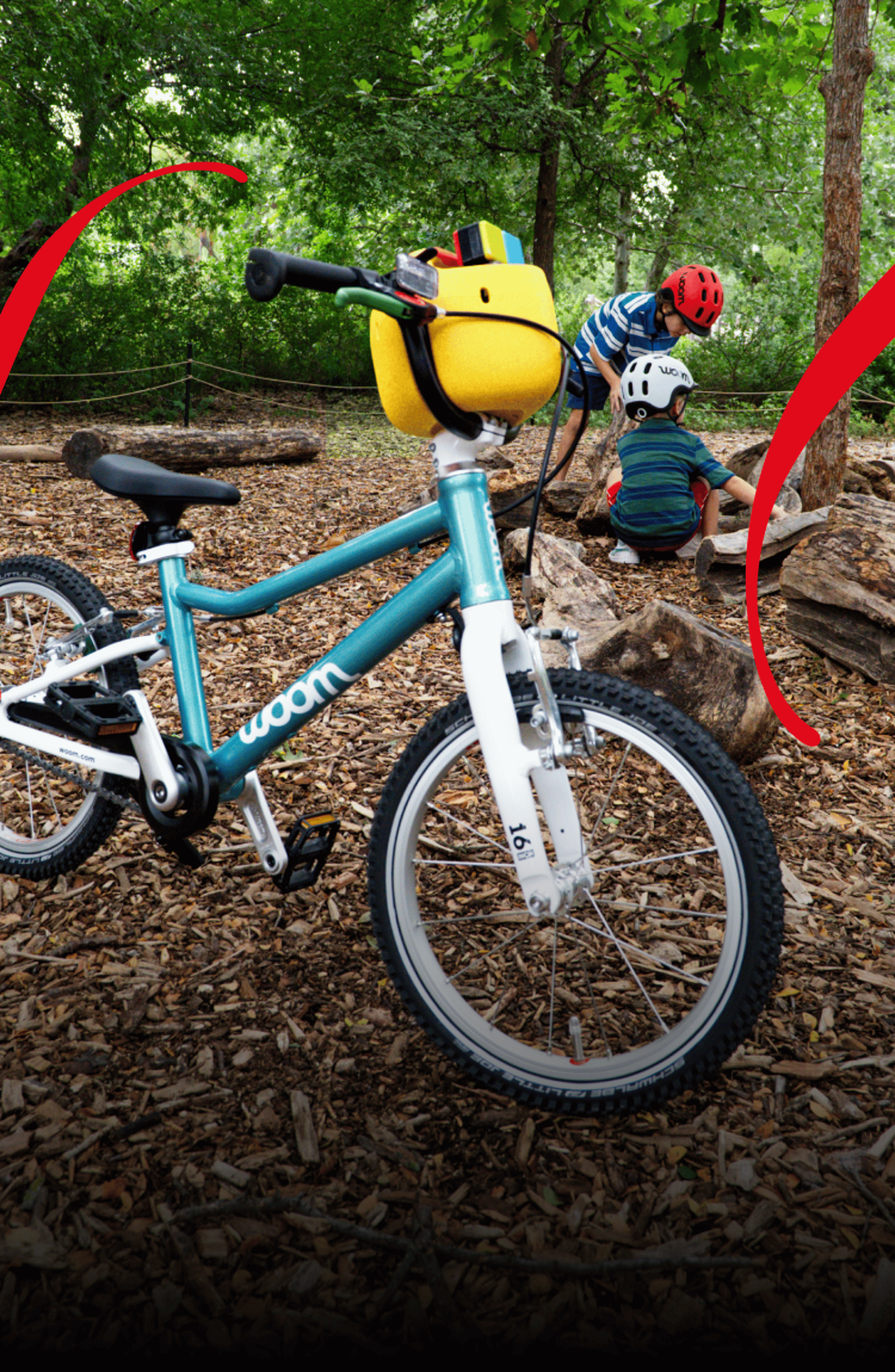 A woom GO in metallic turquoise with a yellow POP bicycle basket on the handlebars stands on a forest floor covered with wood chips, while two children wearing woom READY helmets play in the background.