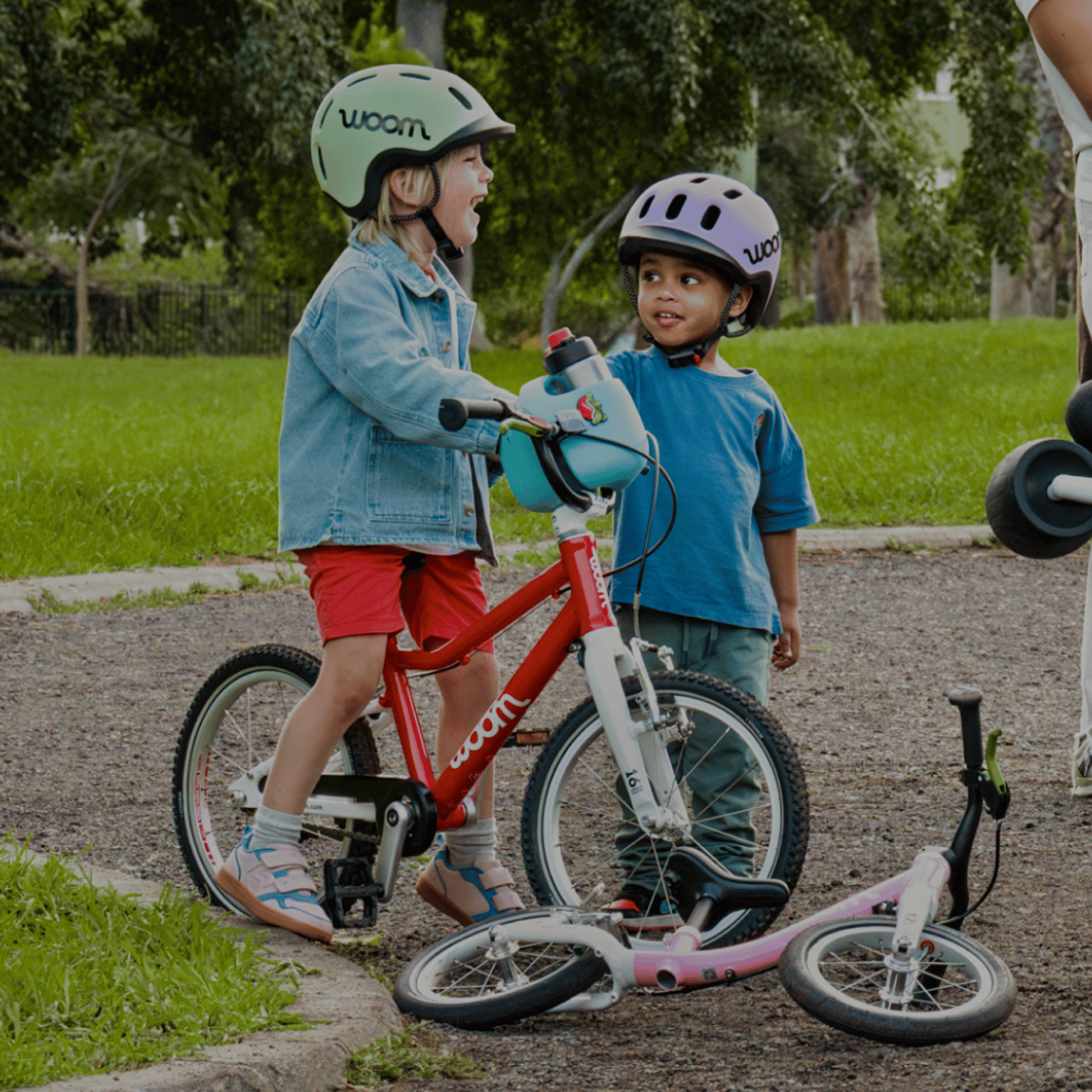 Two kids in READY Helmets chat next to their red and powder pink woom GO bikes.