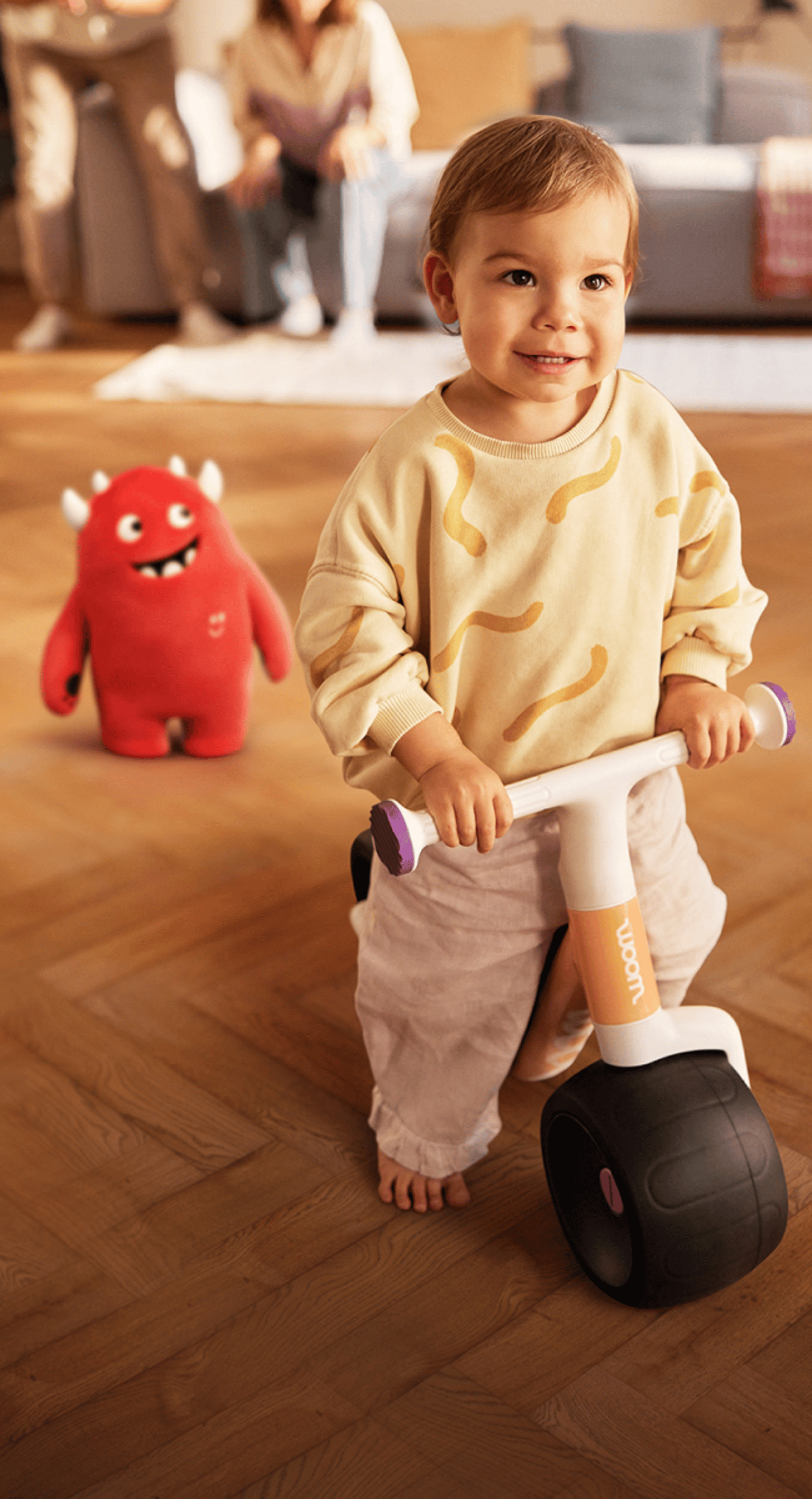 Child sitting on woom WOW balance bike, smiling, parents in the background.