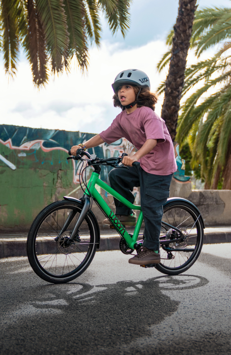 Child wearing a woom READY helmet riding a green woom EXPLORE bike past a graffiti wall.