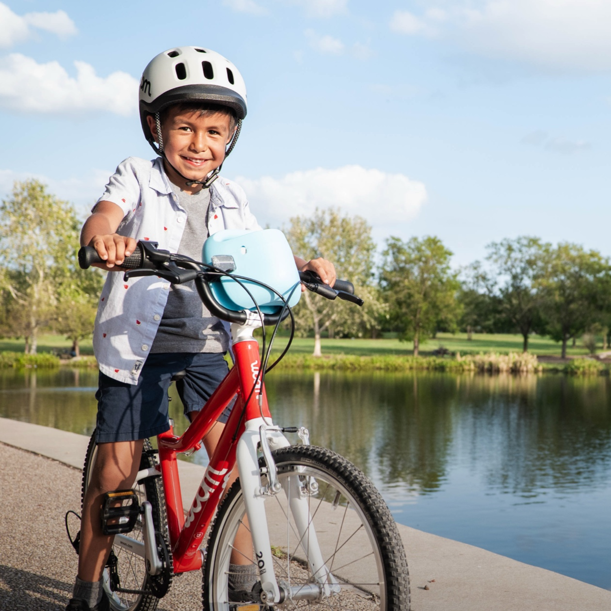     * A boy on a paved path next to a tree-lined lake wearing a pale grey READY Kids’ Bike Helmet and riding a red woom GO bike with a bubblegum blue POP Kids’ Bike Basket attached to the handlebars