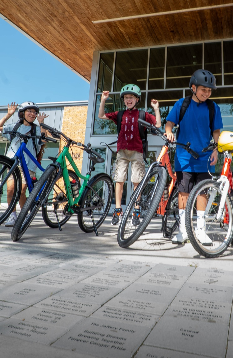 group of kids with woom EXPLORE and woom GO bikes to school while wearing woom READY Kids' Helmets and backpacks standing in front of a school