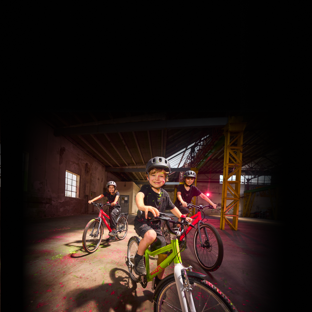 A photo of three kids riding their bikes through a paint-splattered warehouse, highlighting the electric new colors featured on each bike: neon lime woom GO and neon pink woom EXPLORE.