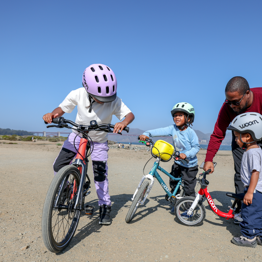 Three kids in READY Helmets prepare to ride their woom bikes on the beach with the help of their dad.