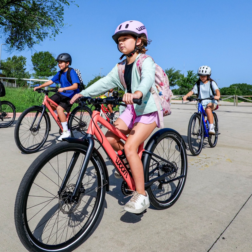 Four kids ride their fully equipped woom bikes through a school parking lot, complete with GLUG water bottles, SNAP click-on fenders, LEGGIE kickstand, and BING bell. Now including BEAM bike lights.