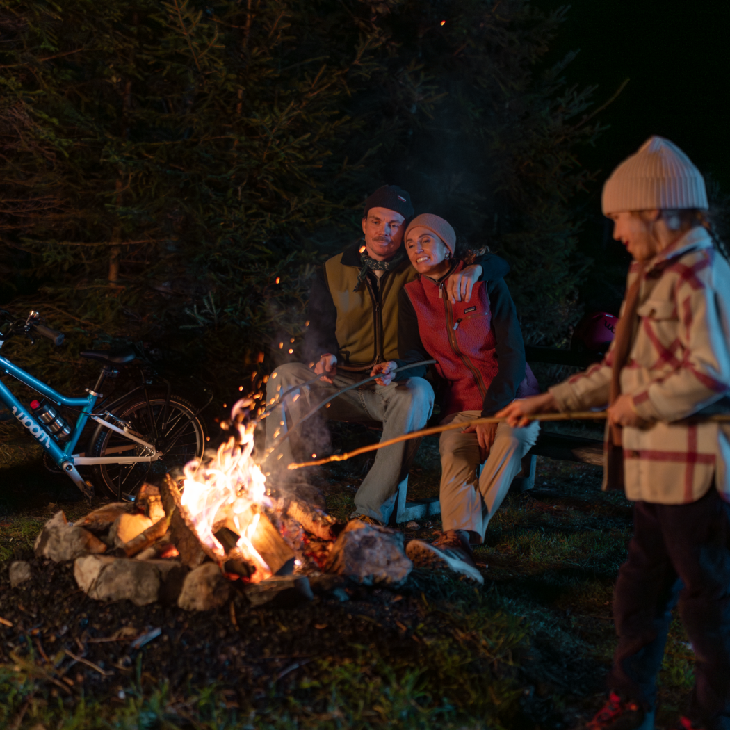 A family gathers around a campfire at night with woom bikes parked at the campsite.