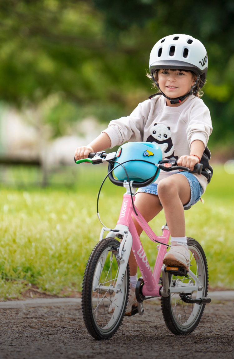 Child in grey READY Kids helmet on powder pink woom GO bike; a woman cheers with open arms on a park path. 