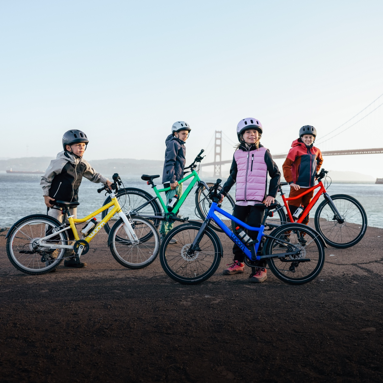 Kids pose with their colorful woom GO and EXPLORE bikes in front of the Golden Gate Bridge on a chilly day.