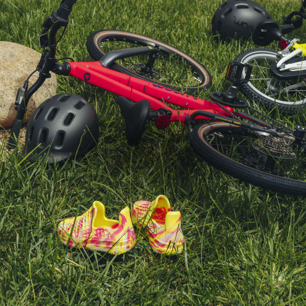 Two NEON woom bikes, a GO and an EXPLORE, lay in the grass next to a pair of neon swirl Lusso Cloud shoes.