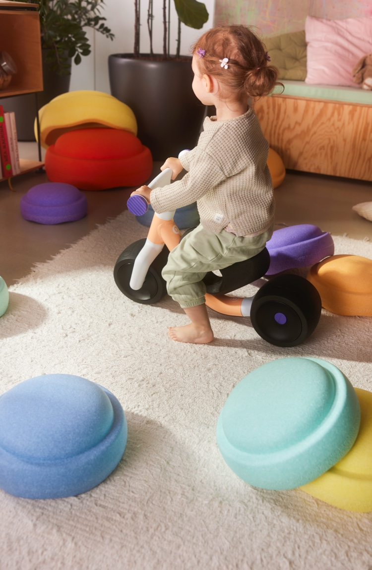 A young child rides a woom WOW in pop peach while surrounded by Stapelstein products