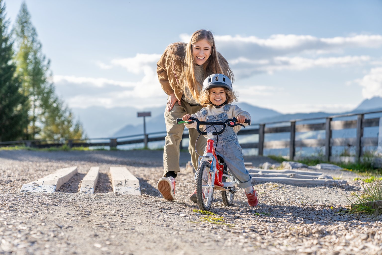 A smiling child wearing a grey woom READY helmet red woom GO bike on a gravel path, with an adult walking closely behind her.