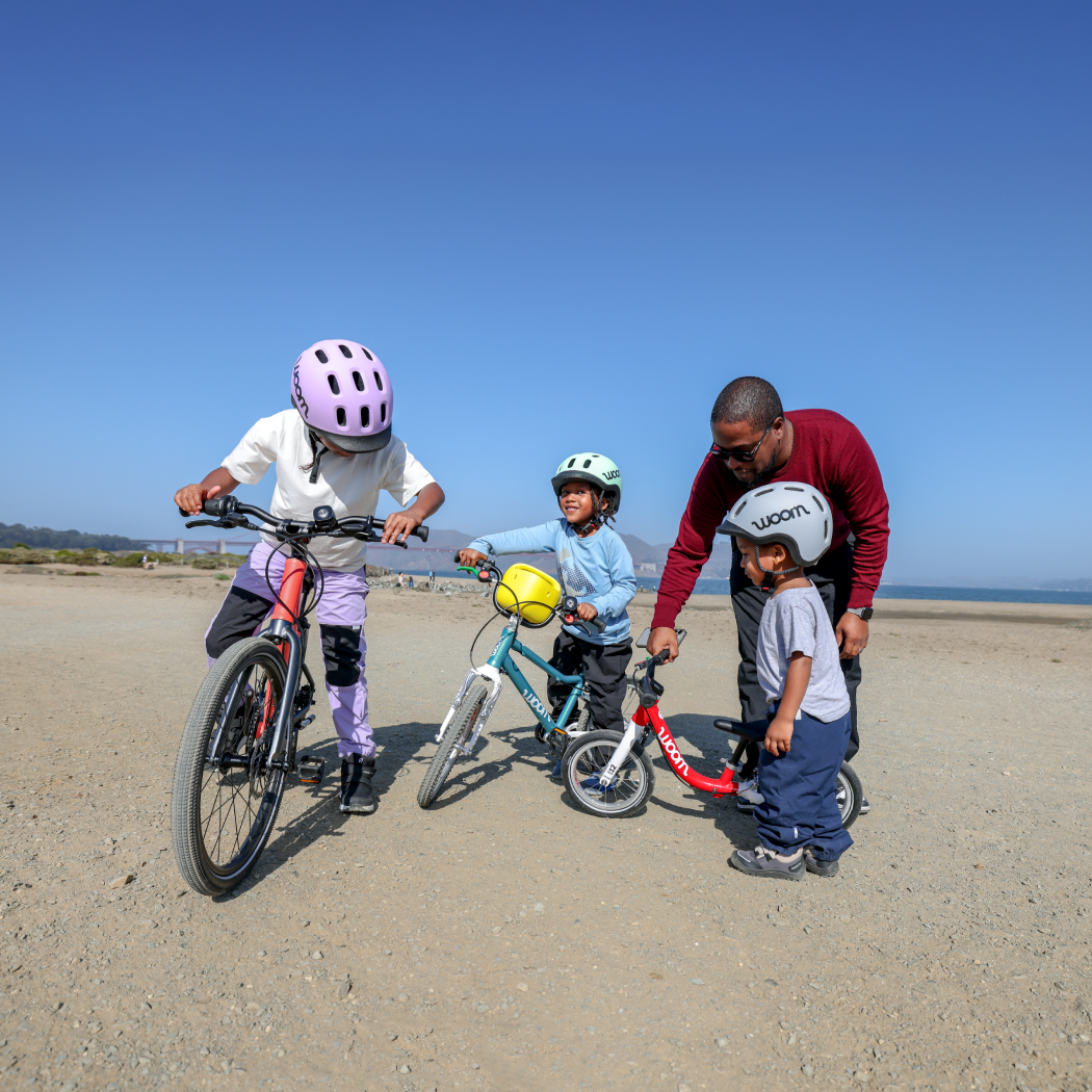 Three kids in READY Helmets prepare to ride their woom bikes on the beach with the help of their dad.