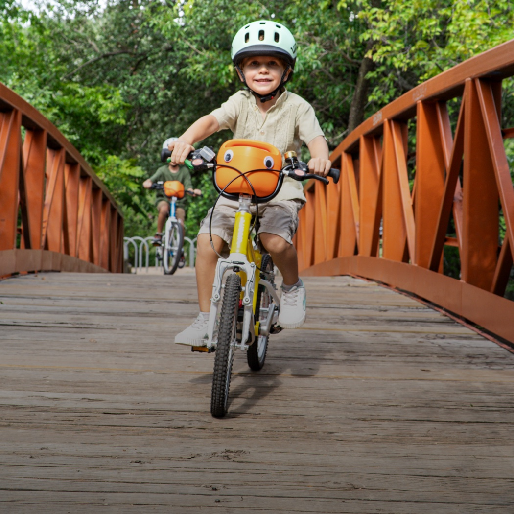 A boy rides a yellow woom GO bike with new orange POP bike basket attached. He is crossing a bridge while wearing a READY helmet, and another young boy rides several paces behind.