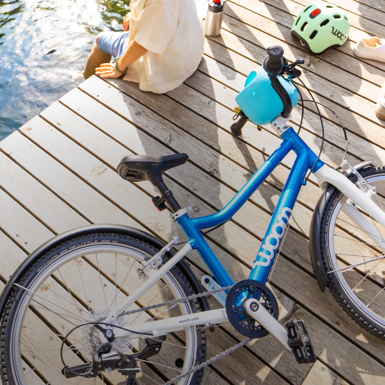 Top-down view of a young boy sitting on the edge of a dock with his feet in the water, next to a metallic blue woom GO 4 with a blue POP Bike Basket attached to the handlebars.