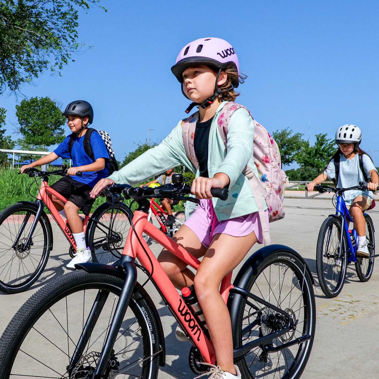 A group of young bike riders steer their woom EXPLORE bikes through a school parking lot. They are wearing woom READY helmets and their magnetic blue and neon coral woom EXPLORE bikes stand out against a bright blue sky.