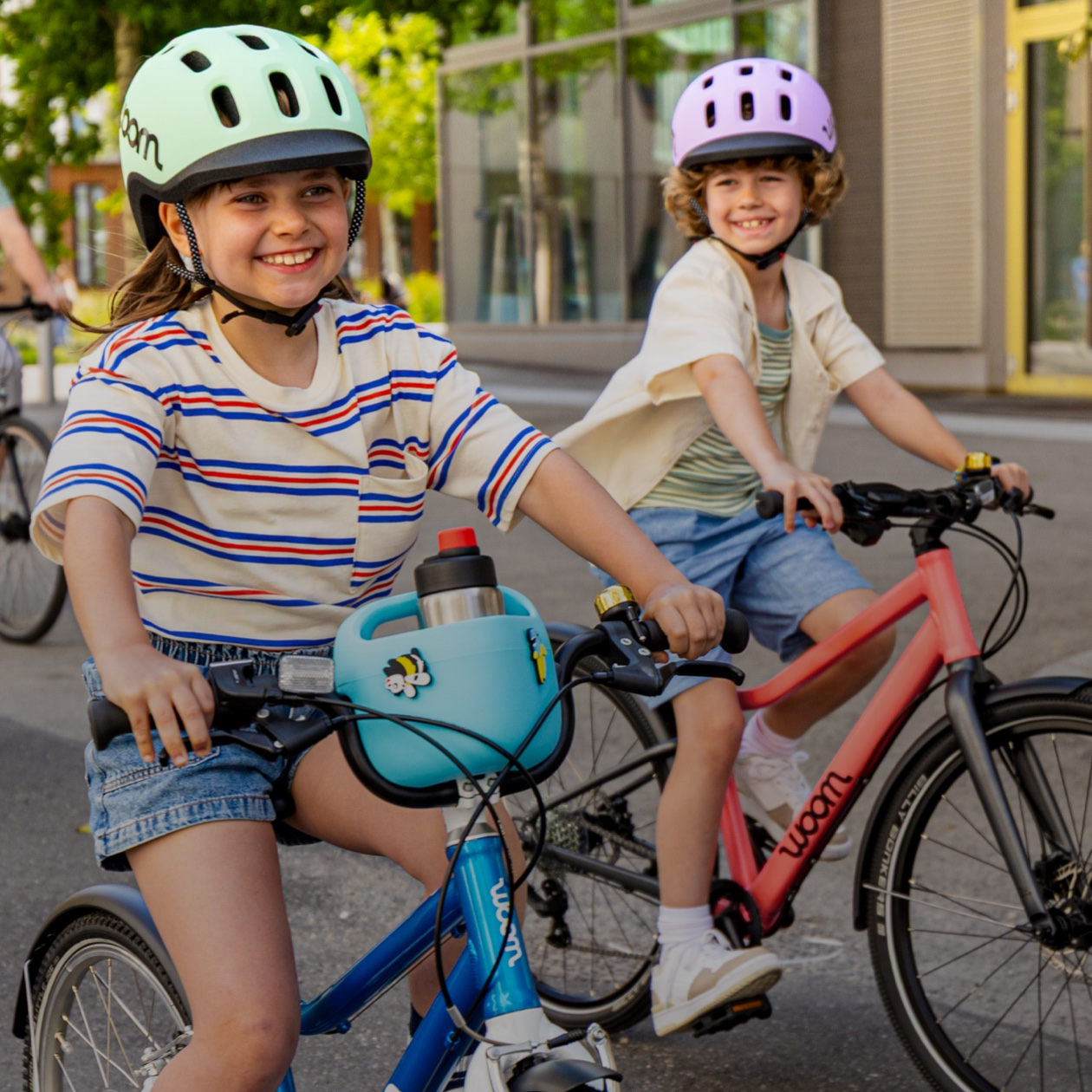 Two kids in woom READY Helmets ride their woom bikes, one with a POP Kid’s Bike Basket and GLUG Stainless Steel Water Bottle, down an empty street on a sunny day.