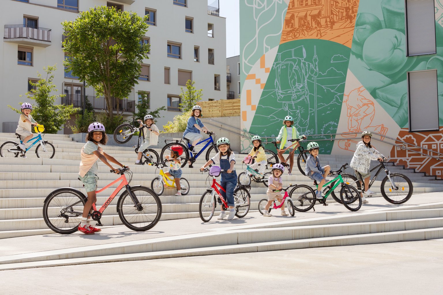 Flera barn med READY-hjälmar på huvudet står med sina woom-cyklar på en trappa i urban miljö.