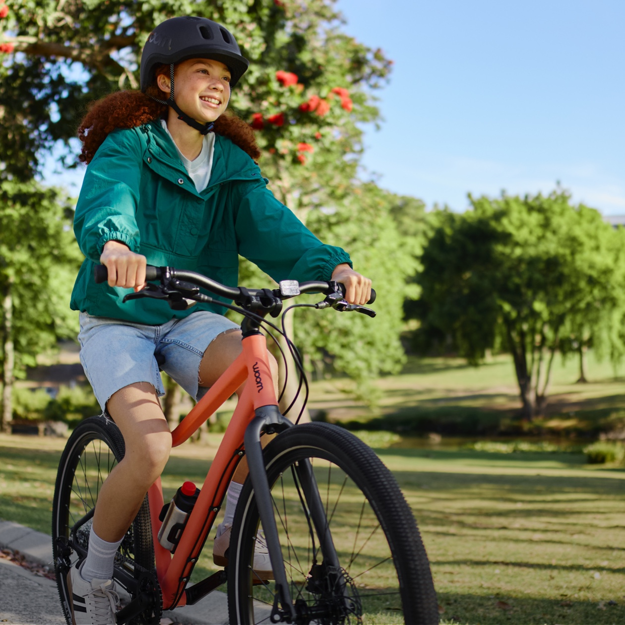 Girl in a black READY helmet with curly red hair, teal windbreaker, and denim shorts rides a neon coral woom EXPLORE bike into the left side of the image. She is in a vibrant, tree-lined park on a sunny day.