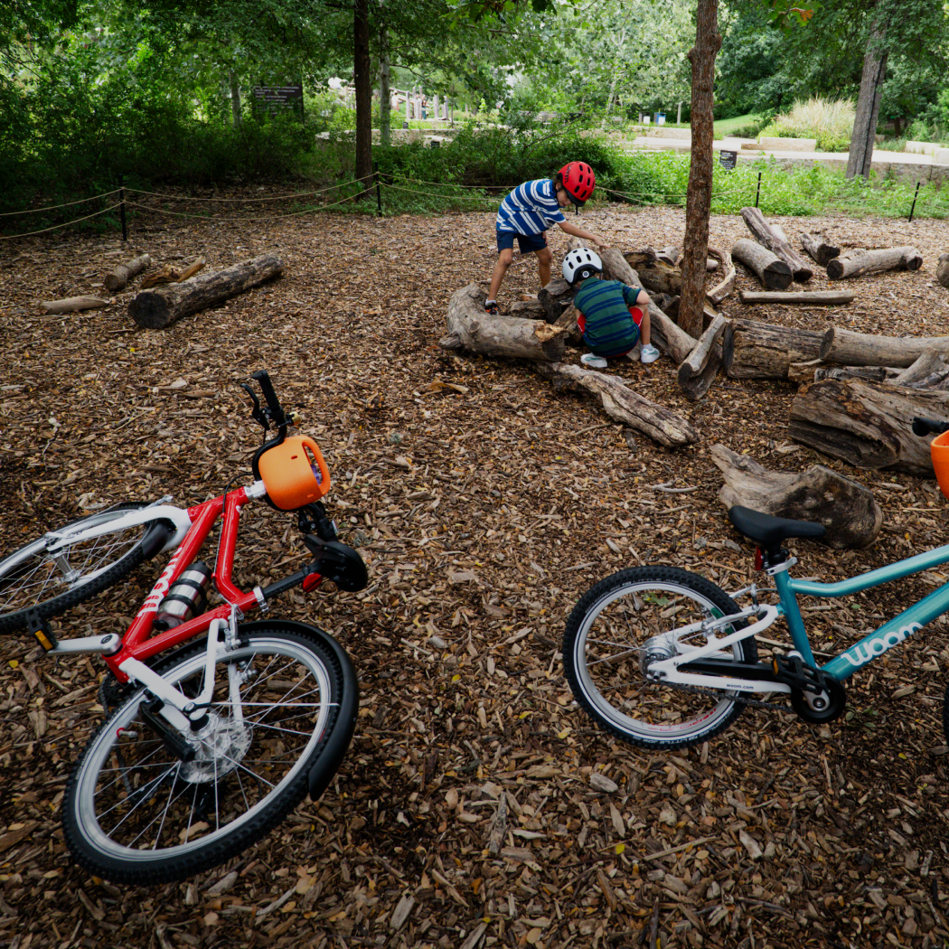 Two boys explore logs in a wooded park area while wearing their woom READY Helmets, with their woom GO bikes laying on the ground nearby.
