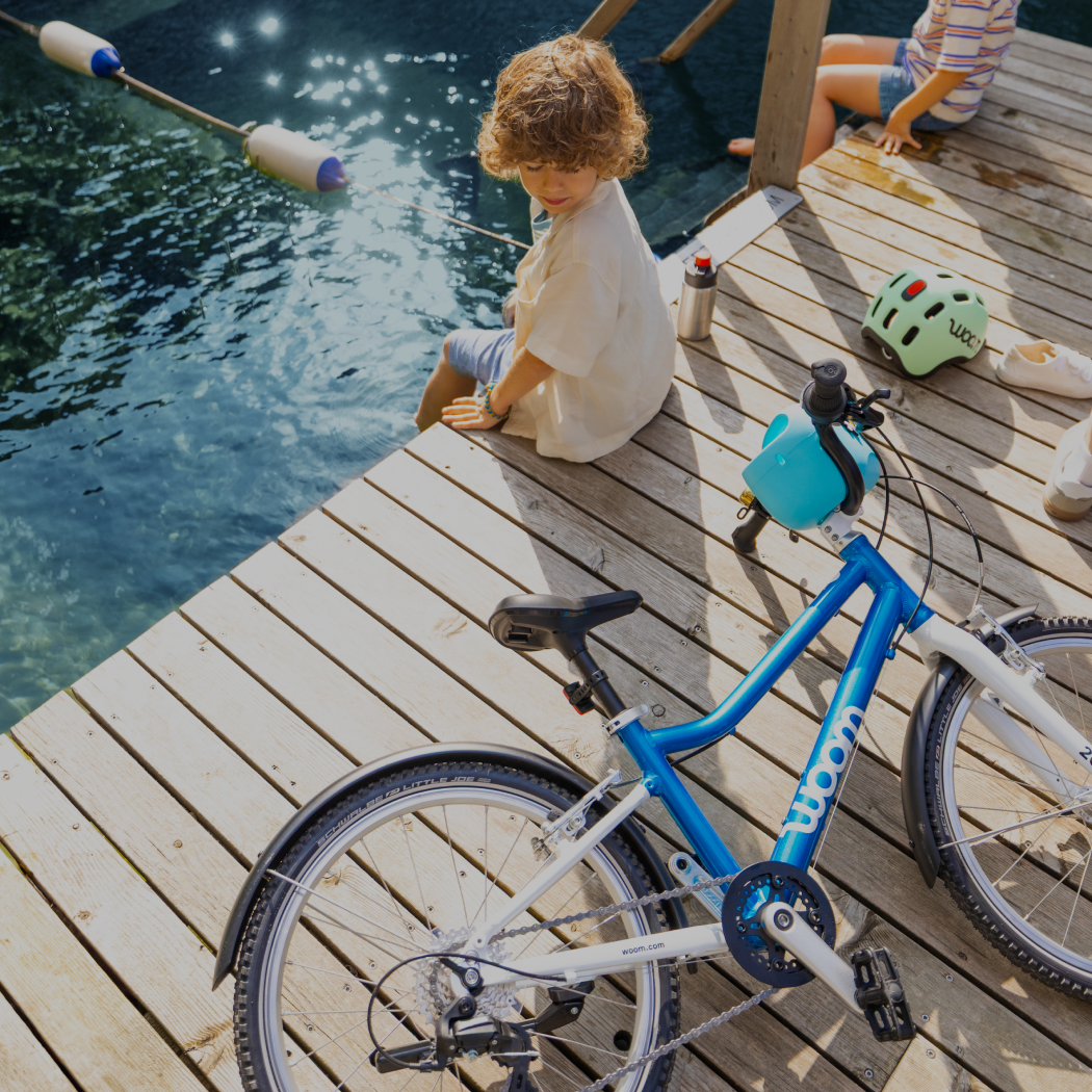An overhead image of a boy sitting on a dock and dangling his feet into blue water. He’s looking at his metallic blue woom GO 4 lying nearby alongside a GLUG Water Bottle and READY Kid’s Helmet.