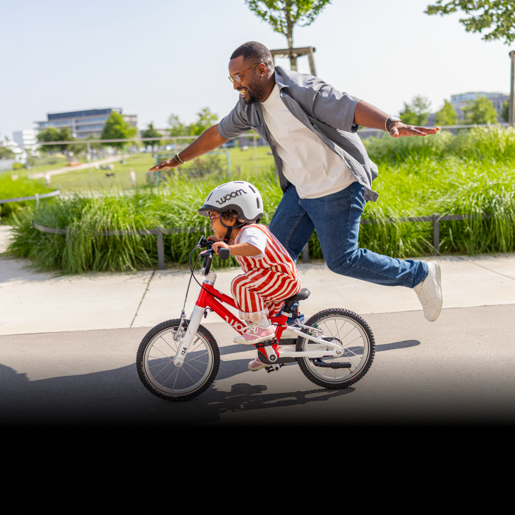 A father runs cheerfully next to his young son as he successfully pedals his red woom GO 2 down an empty street next to a park.